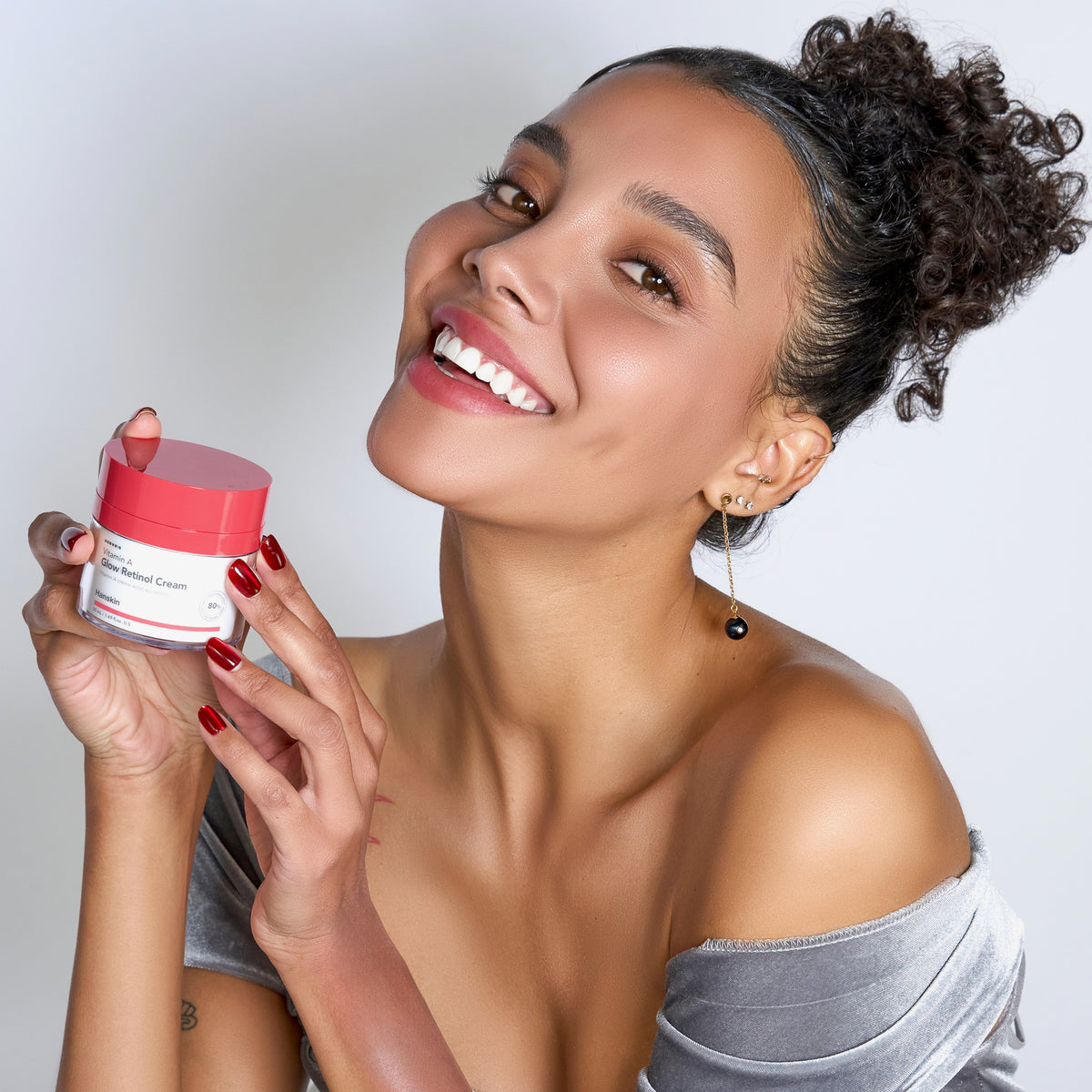 Eine lächelnde Frau mit hochgestecktem Lockenhaar, einem grauen Samtoberteil  hält einen Tiegel Vitamin A Glow Retinol Cream.