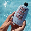 Hände halten eine Flasche Sun Tan Sealer aus der PURISH Summer Bag 2025 an einem Pool, im Hintergrund blaues Wasser.