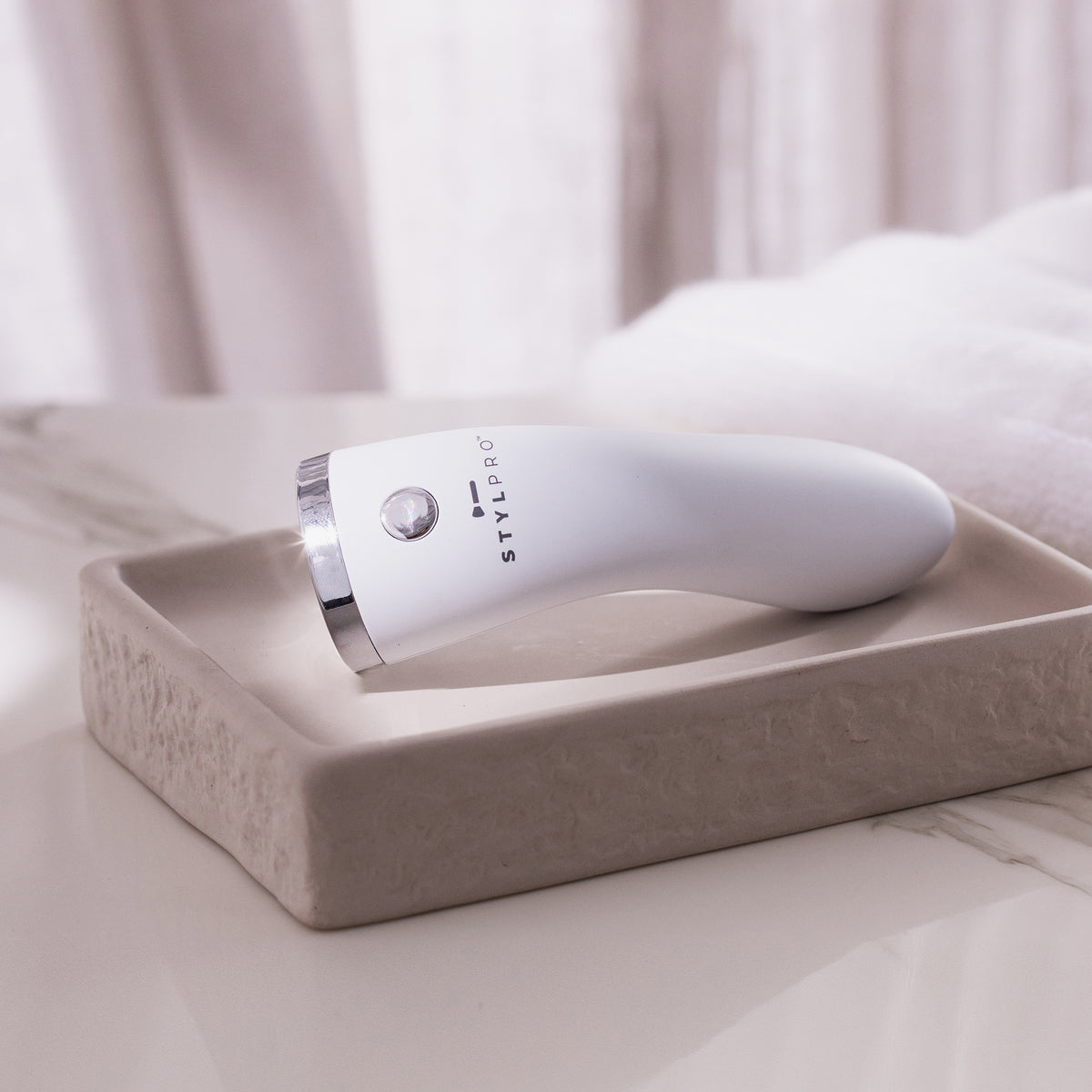 Der STYLPRO Brightspot Anti-Blemish LED Wand, ein elegantes, weißes Handgerät für die Schönheitspflege, steht auf einem beigen Tablett auf einer Marmor-Arbeitsplatte.