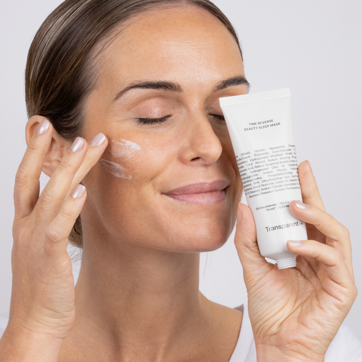 Eine Frau mit geschlossenen Augen trägt die Maske auf ihrer rechten Wange auf und hält das Produkt in ihrer anderen Hand vor das Gesicht.