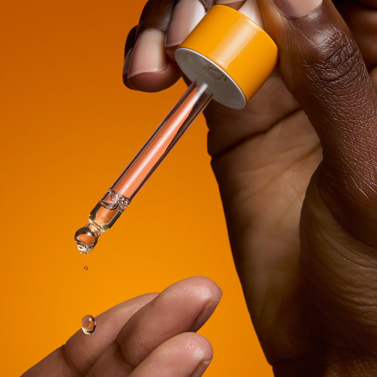 Eine Hand hält eine Pipette vor einem orangefarbenen Hintergrund und gibt das Serum auf die Fingerspitzen ab.