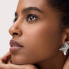 Eine Frau mit glatter Haut und strukturierten Ohrringen blickt nach oben und betont ihre Wimpern mit dem TUBING Stay In Place Volume Mascara 010.