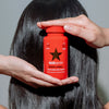 Hände halten eine rote Flasche HAIRtamin Advanced Formula 1 Month vor langes, glattes, glänzendes dunkles Haar.