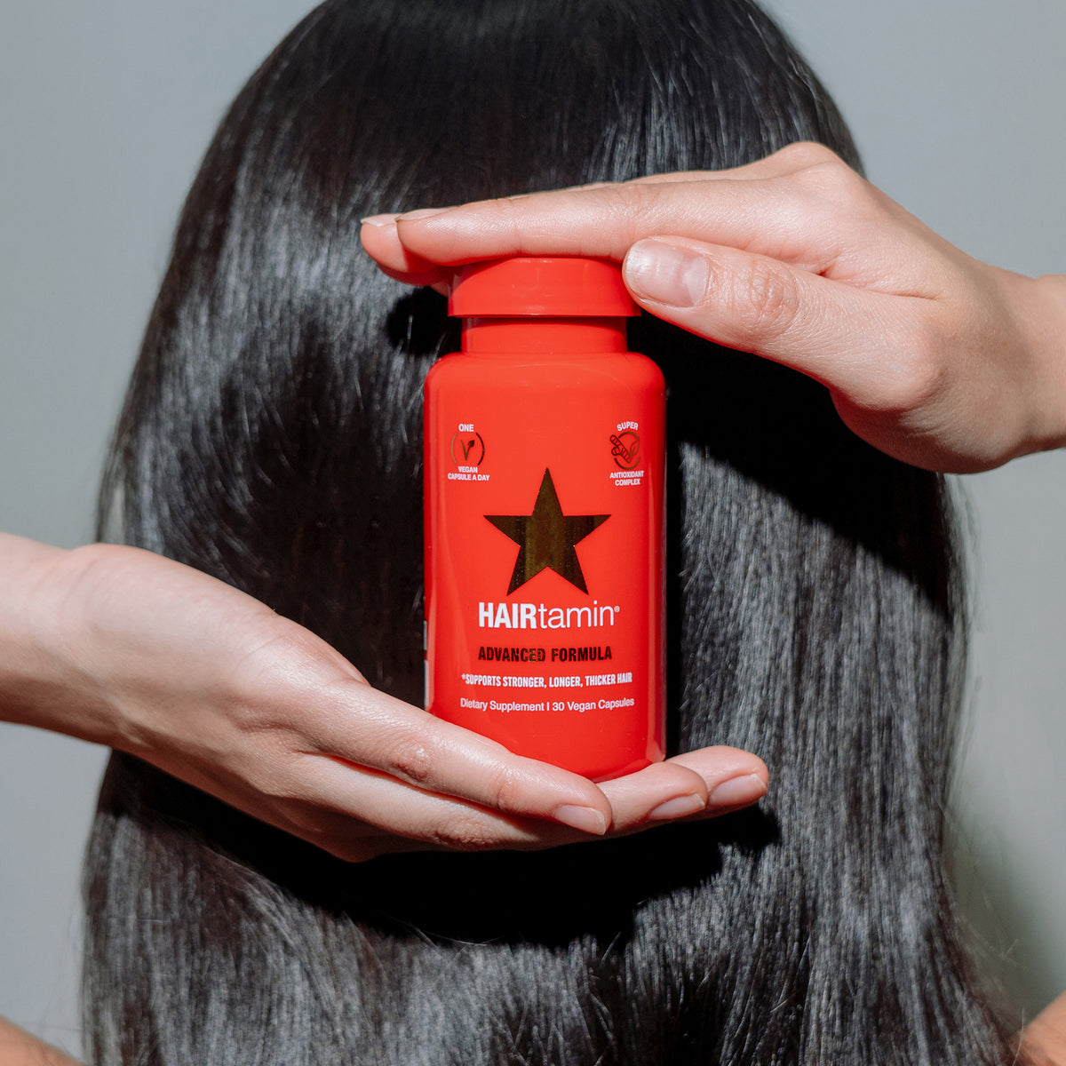 Hände halten eine rote Flasche HAIRtamin Advanced Formula 1 Month vor langes, glattes, glänzendes dunkles Haar.
