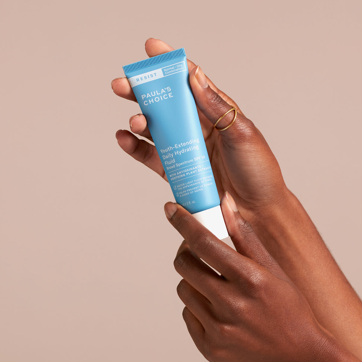 Eine Hand hält eine blaue Tube Youth-Extending Daily Hydrating Fluid SPF 50 vor einem beigen Hintergrund.