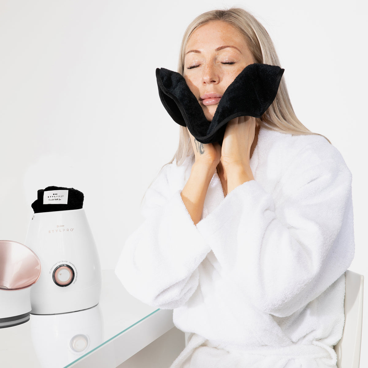 Frau im weißen Bademantel hält ein schwarzes Handtuch an ihr Gesicht. Daneben steht der Gesichtsdampfer in Roségold auf einem Glastisch.
