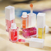 Mehrere Flaschen Glossin‘ Glow Tinted Lip Oil im Freien auf einer hellgelben Oberfläche.