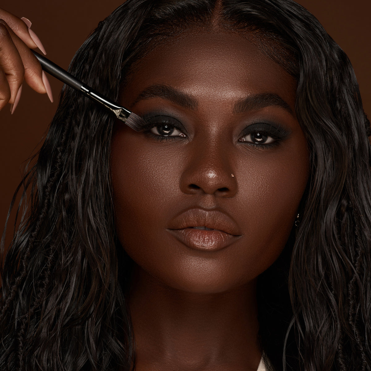 Eine Frau mit langem, dunklem Haar und dunklem Teint trägt mit einem Make-up-Pinsel Lidschatten aus der Groundwork Palette – Defining Neutrals auf.