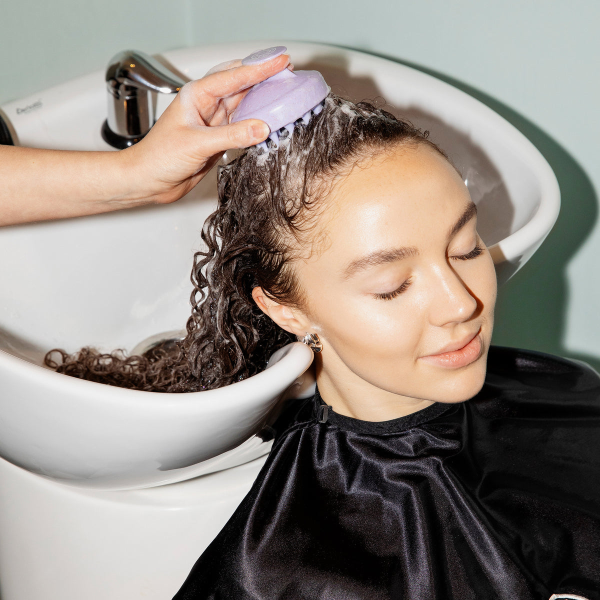 Die lockigen langen Haare einer Frau werden am Waschbecken eines Friseursalons gewaschen und die Kopfhaut mit der maria nila Stimulating Scalp Brush massiert.