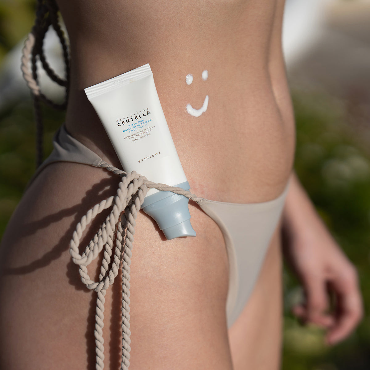 Die PURISH Summer Bag 2025 – Basic Routine sitzt auf einem Bikini, während ein mit Lotion aufgemaltes Smiley-Gesicht den Bauch der Person ziert.