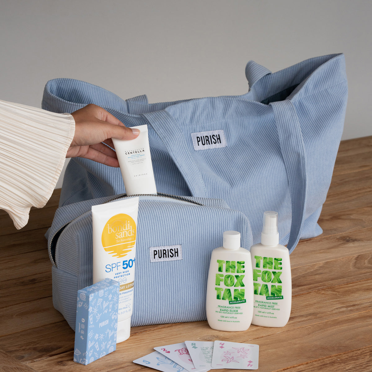Eine Hand packt Hautpflegeprodukte und Karten in die PURISH Summer Bag 2025 – Basic Routine, die auf einer Holzoberfläche steht.