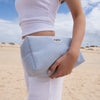 Person in weißer Kleidung hält eine hellblaue PURISH Summer Bag 2025 – Basic Routine an einem Sandstrand mit bewölktem Himmel.