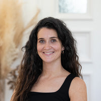 Porträt von Anne-Laure mit dunkelbruanen Wellen, Goldohrringen und schwarzem Oberteil vor hellem Hintergrund.