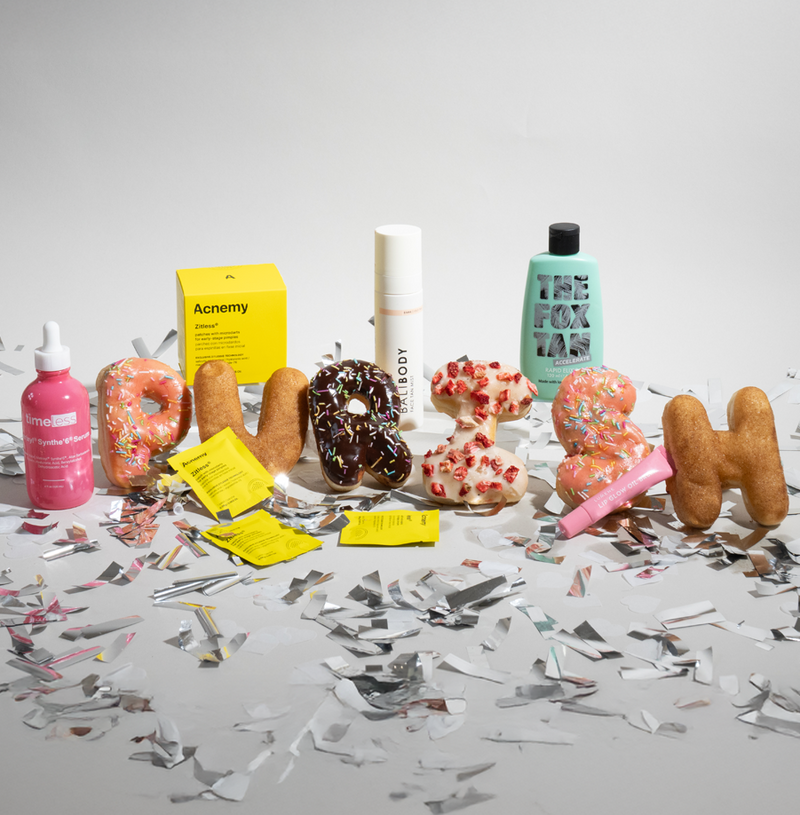 Donuts formen das Wort ‚PURISH‘, umgeben von Kosmetikprodukten und silbernem Konfetti auf weißem Hintergrund.