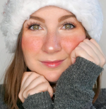Junge Frau mit Sommersprossen, rosa Wangen und glänzenden Lippen trägt eine weiße Mütze und grauen Pulli, blickt freundlich in die Kamera.