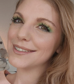 Lächelnde Frau mit grünem Augen-Make-up, langen Wimpern und nudefarbenem Lippenstift vor hellem Hintergrund.