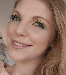 Lächelnde Frau mit grünem Augen-Make-up, langen Wimpern und nudefarbenem Lippenstift vor hellem Hintergrund.