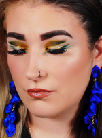 Frau mit geschlossenen Augen, auffälligem gold-grünem Augen-Make-up, blauen Statement-Ohrringen und Nasenring.