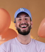 Lachender Mann mit Bart, hellblauem LA-Cap und rosa Shirt steht vor orangem Hintergrund mit Ballons.