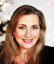 Frau mit langen, dunkelblonden Haaren, rosa Lippenstift und Ohrringen lächelt in die Kamera vor unscharfem, hellem Hintergrund.