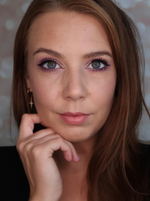Frau mit offenem braunen Haar, pinkem Lidschatten und Eyeliner schaut direkt in die Kamera und stützt das Kinn auf die Hand.
