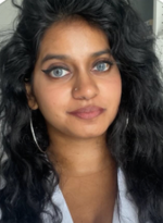 Frau mit langen schwarzen Locken und blauen Augen, auffälligem Eyeliner und silbernen Creolen blickt ruhig in die Kamera, im Hintergrund ein Zimmer.