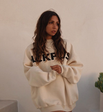 Frau mit langen braunen Haaren trägt beigen Oversize-Pullover mit schwarzem Schriftzug und blickt ernst zur Seite.