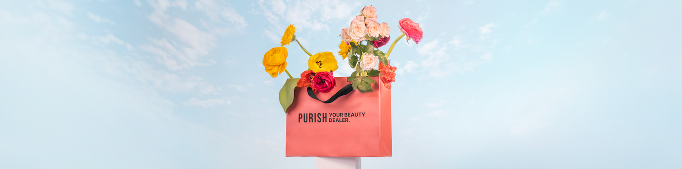 Blumen in leuchtenden Farben stecken in einer rosa Einkaufstasche mit der Aufschrift ‚PURISH YOUR BEAUTY DEALER‘ vor Himmel-Hintergrund.
