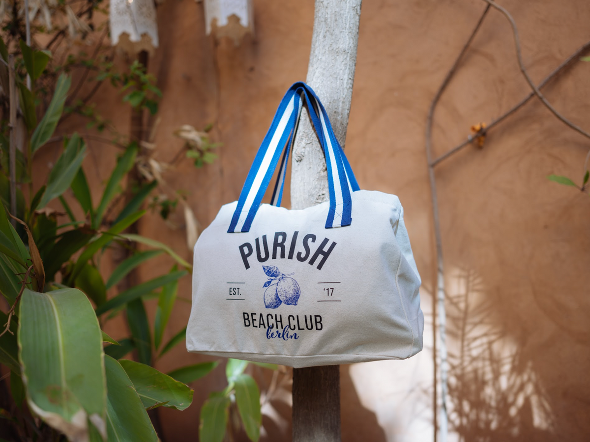 Die erste PURISH Summer Bag mit blauem Aufdruck " PURISH Est. '17 Beach Club Berlin" und Tragegriffen hängt an einem Baum, umgeben von mediterranen Pflanzen.
