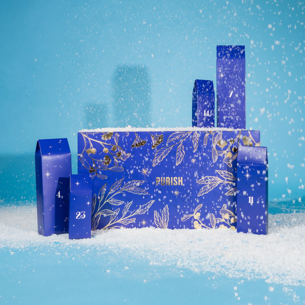 Blauer Box des PURISH Adventskalender mit goldenen Illustrationen und nummerierten Türchenboxen steht im Kunstschnee vor hellblauem Hintergrund.
