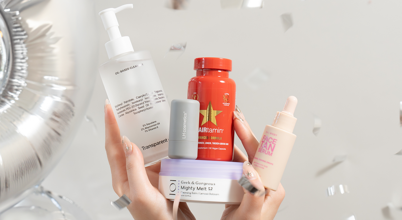 Eine Hand hält mehrere Pflegeprodukte vor silbernem Ballon und Konfetti – darunter Oil-Based Cleanser, Face Tan Serum, HAIRtamin und Mighty Melt.
