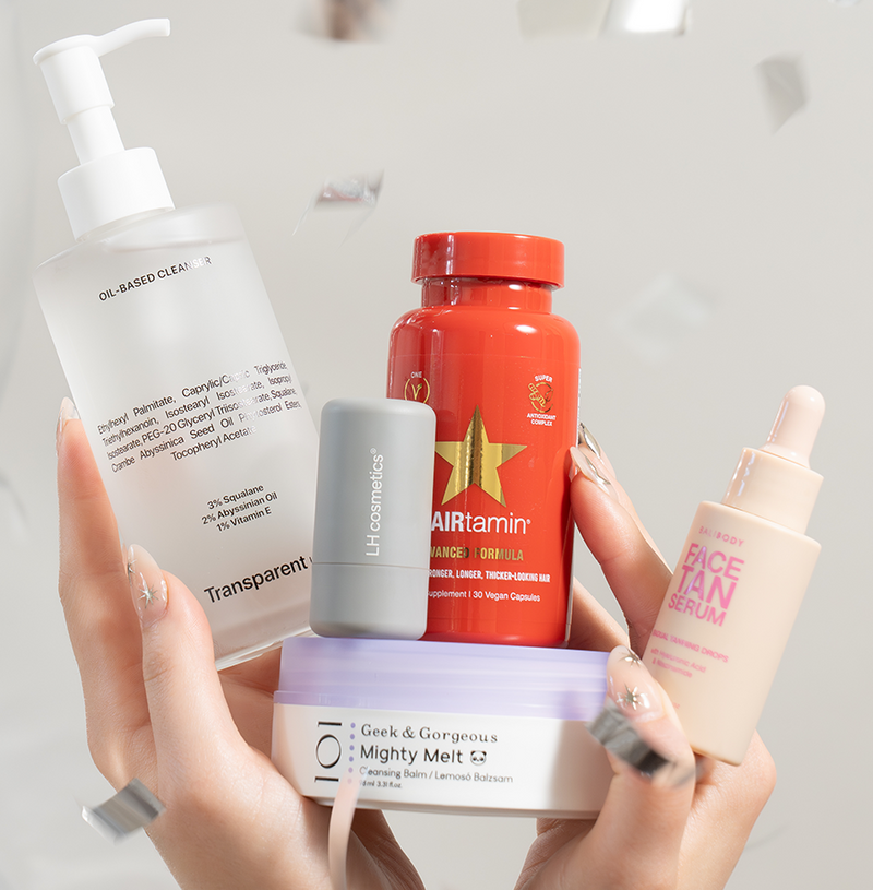 Eine Hand hält mehrere Pflegeprodukte vor silbernem Ballon und Konfetti – darunter Oil-Based Cleanser, Face Tan Serum, HAIRtamin und Mighty Melt.
