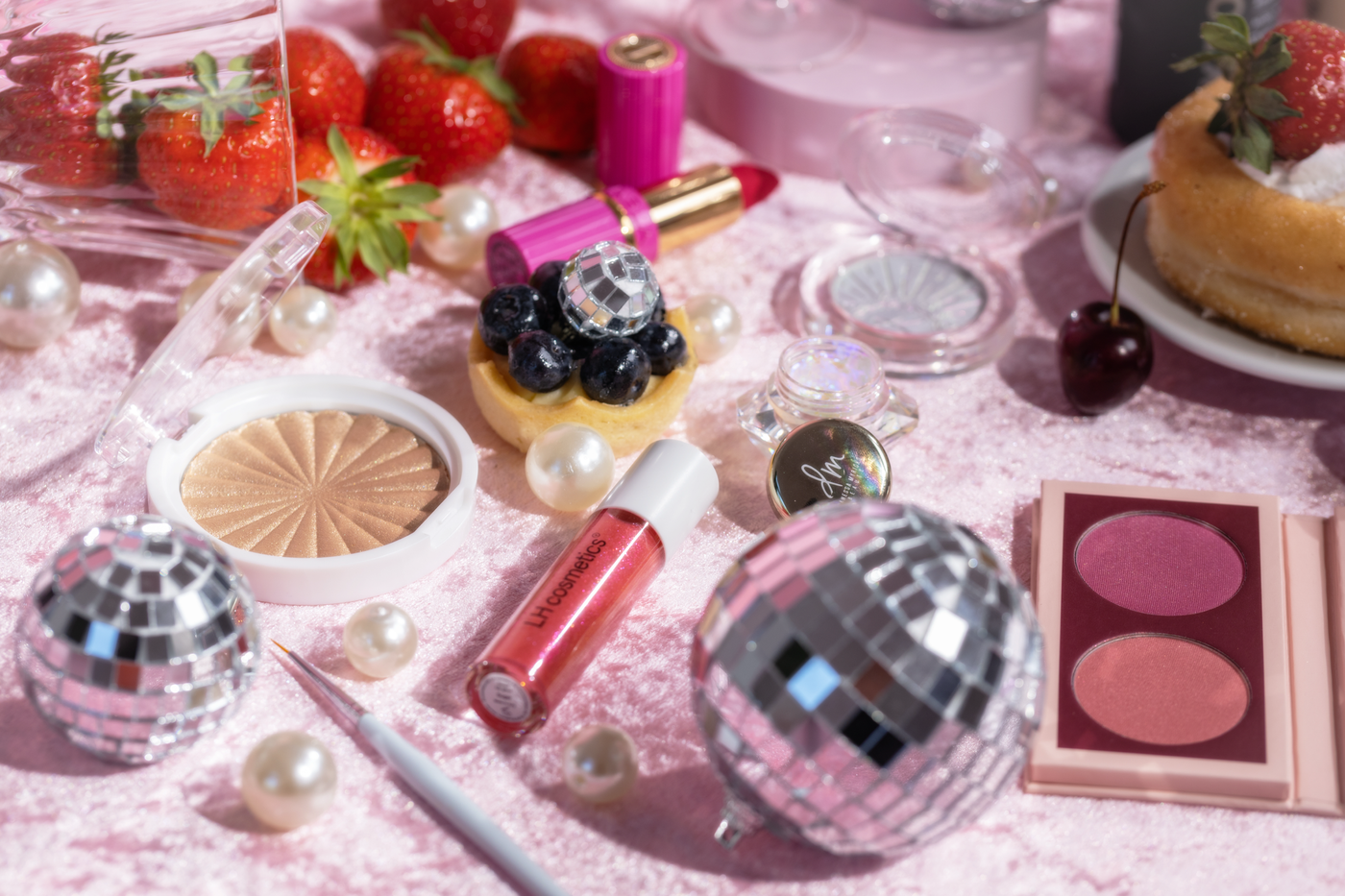 Kosmetikprodukte wie Rouge, Highlighter und Lippenstift liegen auf rosa Samt, umgeben von Erdbeeren, Perlen und Disco-Kugeln.