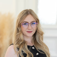 Porträt von Marana mit blonden Haaren, Brille mit violettem Rahmen und schwarzem Oberteil vor hellem Hintergrund.