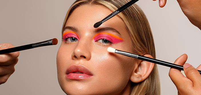Frau mit auffälligem pink-orangefarbenem Augen-Make-up wird mit mehreren Pinseln gleichzeitig geschminkt.