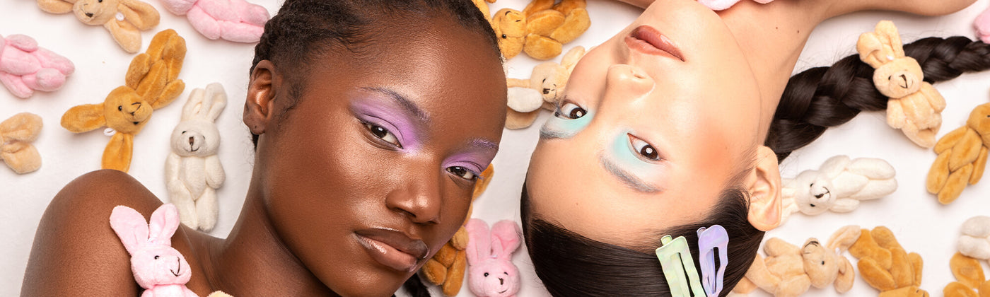Zwei Models mit farbigem Lidschatten liegen zwischen kleinen Plüschhasen auf weißem Hintergrund, ihre Haare sind geflochten.