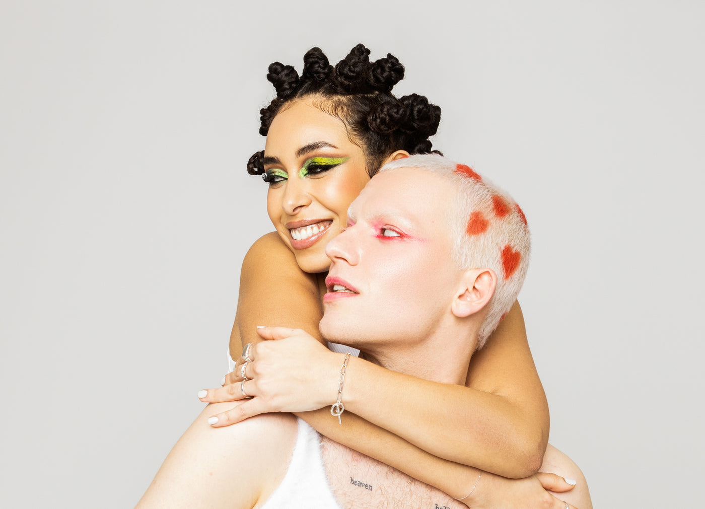 Zwei Models mit auffälligem grünen und roten Make-up umarmen sich und lächeln; eines trägt Bantu-KNots, das andere hat rote Herzchen im weißen kurzen Haar.