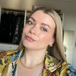 Frau mit glattem blonden Haar und floralem Oberteil posiert lächelnd vor einem offenen Kleiderschrank und einem Spiegel im Hintergrund.