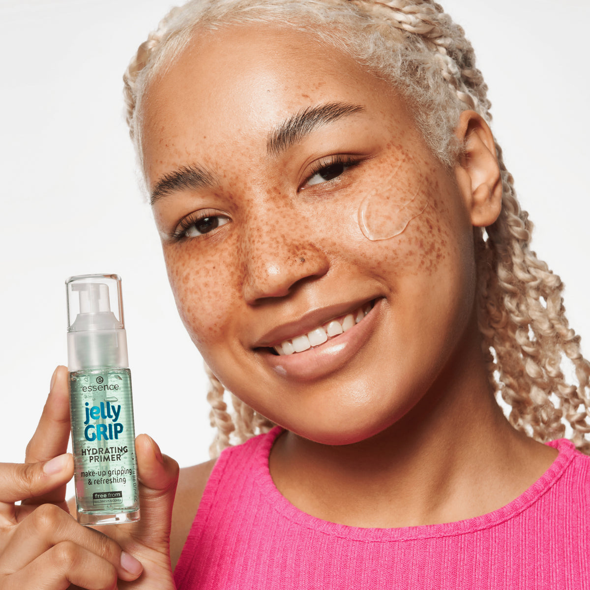 Lächelnde Person mit Sommersprossen in einem rosa Oberteil hält eine Flasche Jelly GRIP HYDRATING PRIMER vor einem weißen Hintergrund.