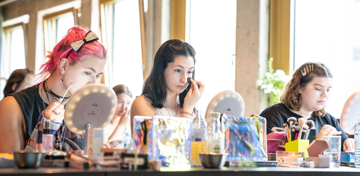 Mehrere Personen sitzen an einem Tisch mit Schminkspiegeln und Pinseln und tragen Make-up auf, in heller, freundlicher Umgebung.