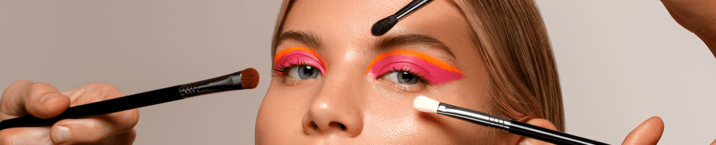 Frau mit auffälligem pink-orangefarbenem Augen-Make-up wird mit mehreren Pinseln gleichzeitig geschminkt.