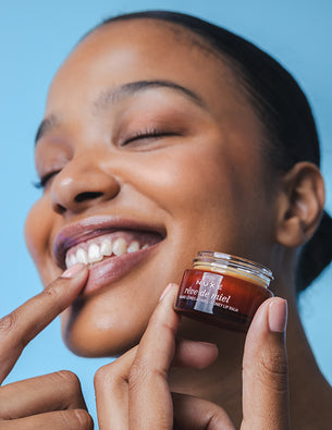 Lächelnde Frau trägt mit dem Finger den NUXE Rêve de Miel Lip Balm auf ihre Lippen auf und hält den braunen Tiegel in der anderen Hand.
