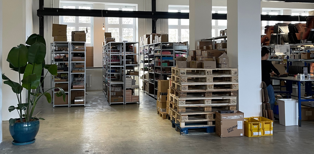 Blick in das neue, größere Lager der PURISH GmbH mit Regalen voller Kosmetikprodukte, Paletten und einer Person beim Verpacken.