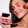 Eine lächelnde Frau trägt mit einem Pinsel das pfirsichfarbene Rouge aus der Duo Dimension Zone Palette von LH Cosmetics auf und hält sie neben ihr Gesicht.