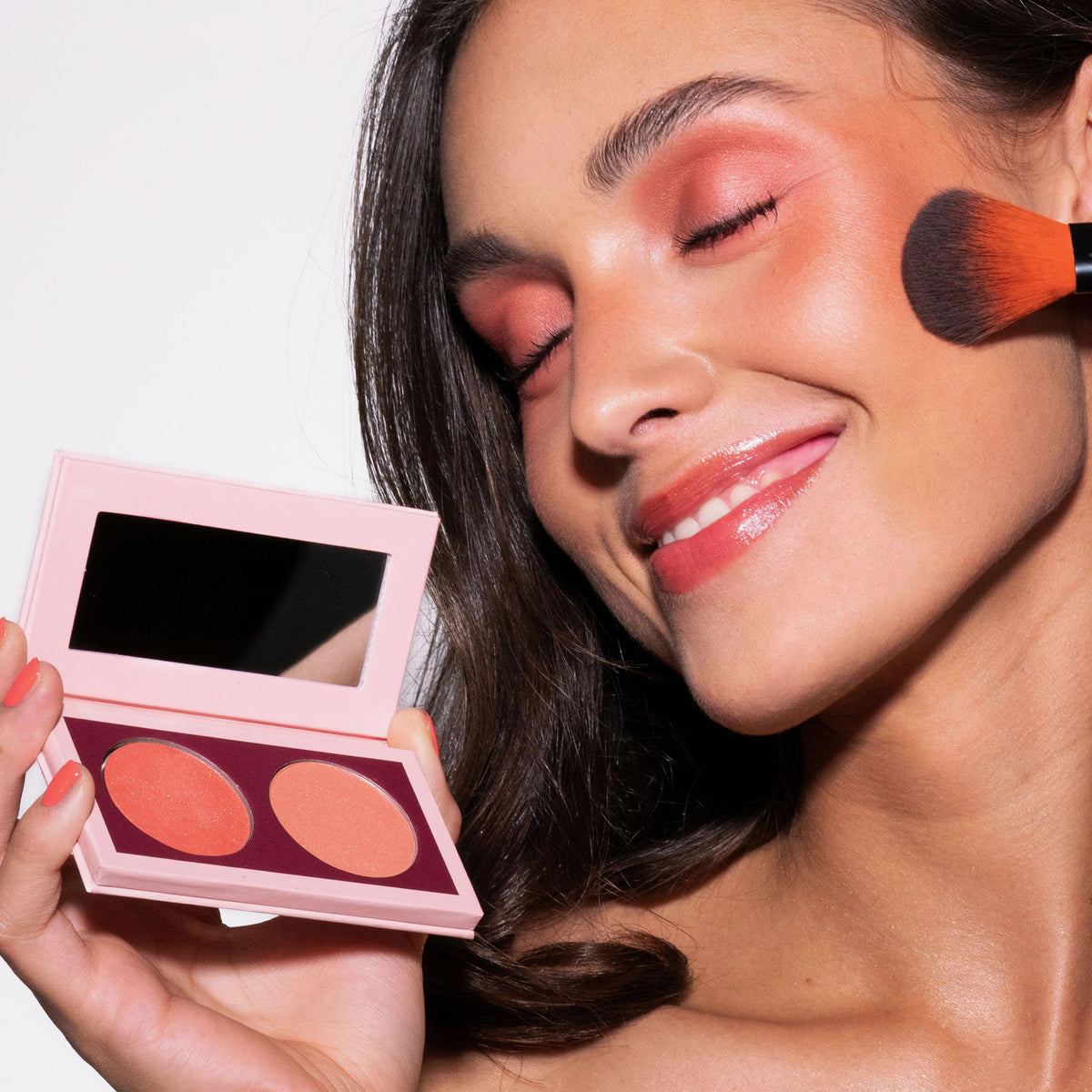 Eine lächelnde Frau trägt mit einem Pinsel das pfirsichfarbene Rouge aus der Duo Dimension Zone Palette von LH Cosmetics auf und hält sie neben ihr Gesicht.