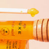 Applikatorstab mit gelbem Lippenbalsam neben einer Tube NUXE Honey Lip Care Rêve de Miel und einem hölzernen Honiglöffel.