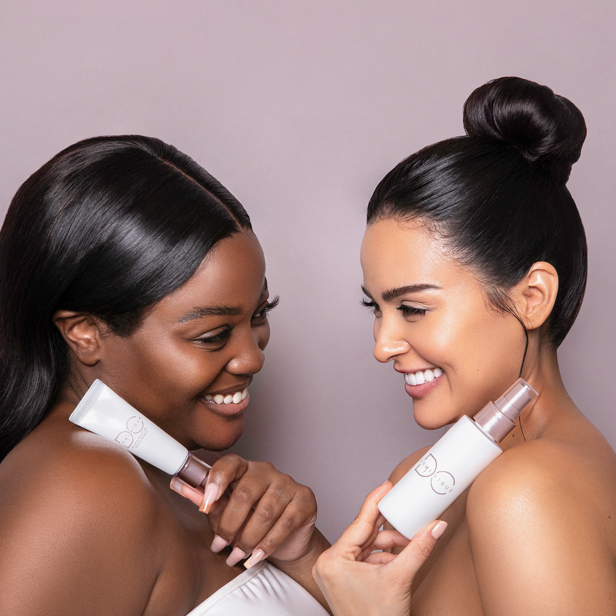 Zwei Frauen lächeln, während sie Ultra Hydrating Fine Mist-Flaschen vor einem hellvioletten Hintergrund halten.