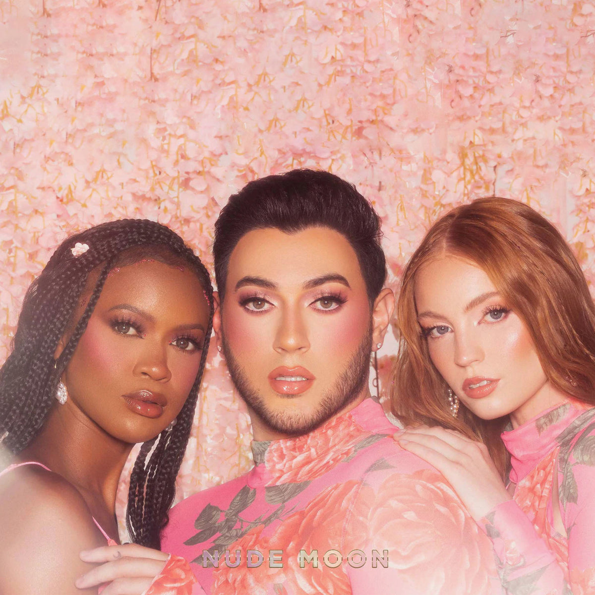 Drei Personen mit glamourösem Make-up posieren vor einem blumigen Hintergrund und präsentieren die Nude Prism Color Palette von Lunar Beauty.