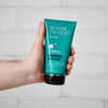 Hand hält eine Tube Men Invigorating Face Scrub vor einem weiß gefliesten Hintergrund.