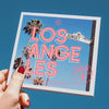 Hand mit roten Nägeln hält die Lost in Los Angeles 16 Color Shadow Palette in Richtung eines blauen Himmels